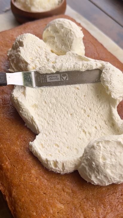 "Dairy-free whipped cream being spread on top of a baked vegan coconut cake with a spatula"