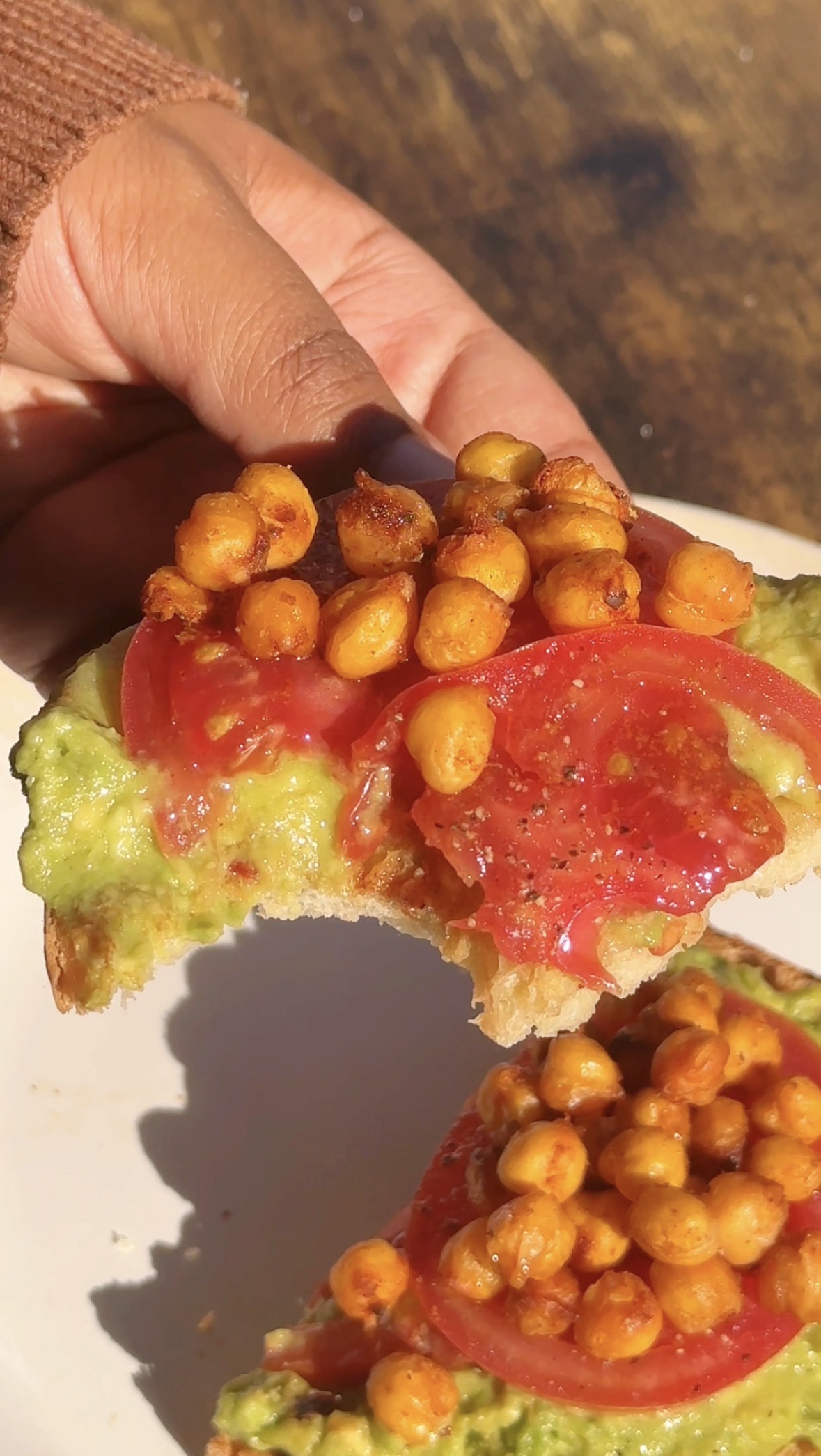 A hand holding a bitten slice of Cajun avocado toast showing crispy chickpeas, tomato, and mashed avocado on sourdough