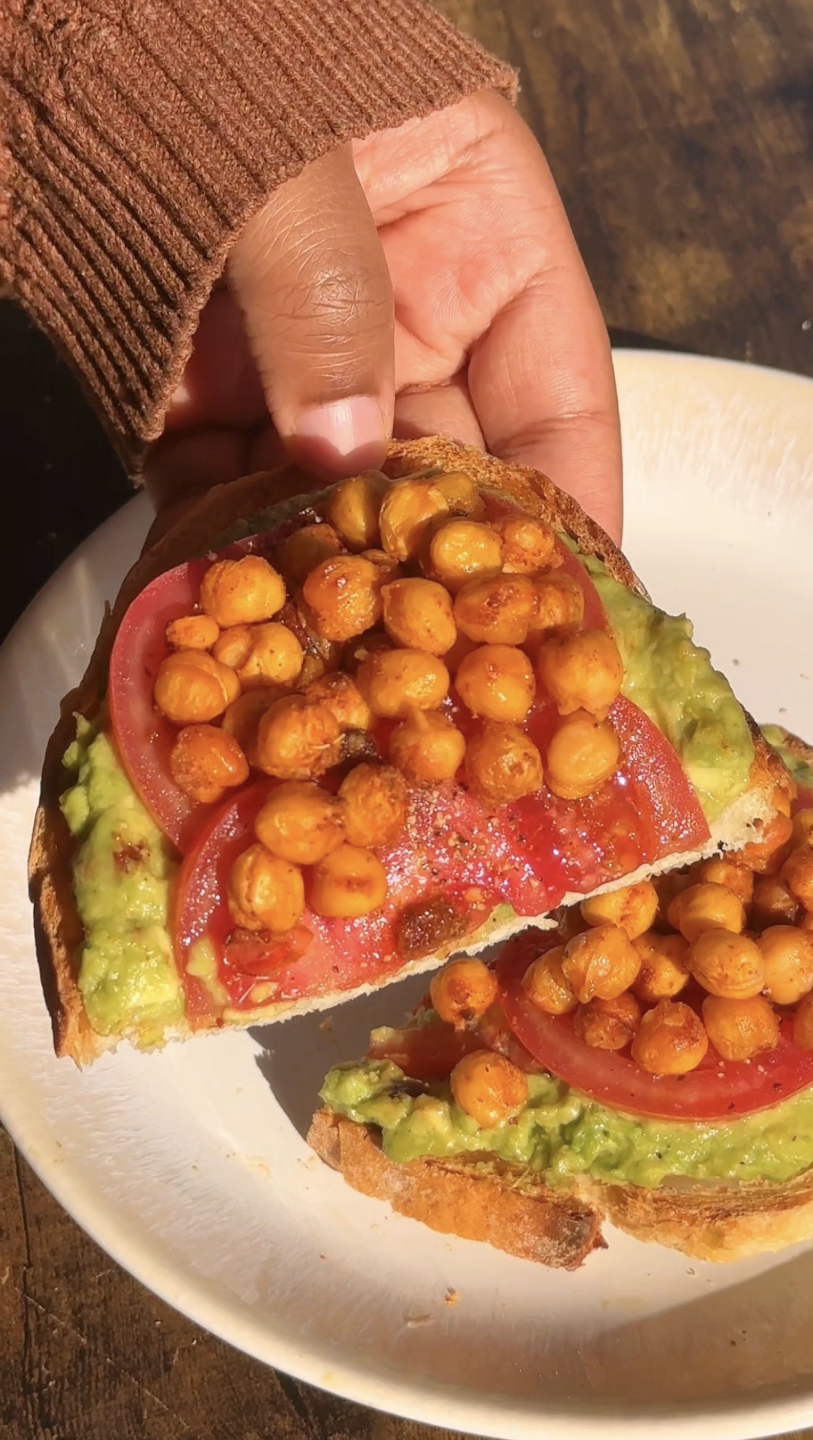 A hand lifting a slice of Cajun avocado toast with crispy chickpeas and tomato, second slice visible on the plate below