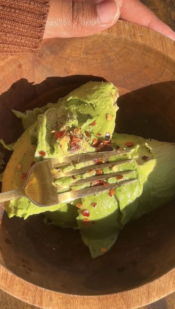 Mashing a ripe avocado with a fork in a wooden bowl with red pepper flakes and seasoning