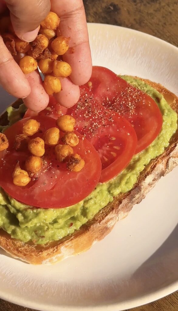 A hand sprinkling crispy Cajun chickpeas onto avocado toast with sliced tomatoes on sourdough